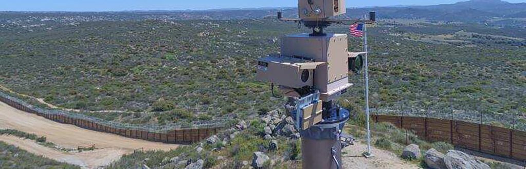 A thin tower covered with various electronic sensors sits in the foreground on a hill with the U.S.-Mexico border wall stretching from left to right in the background.