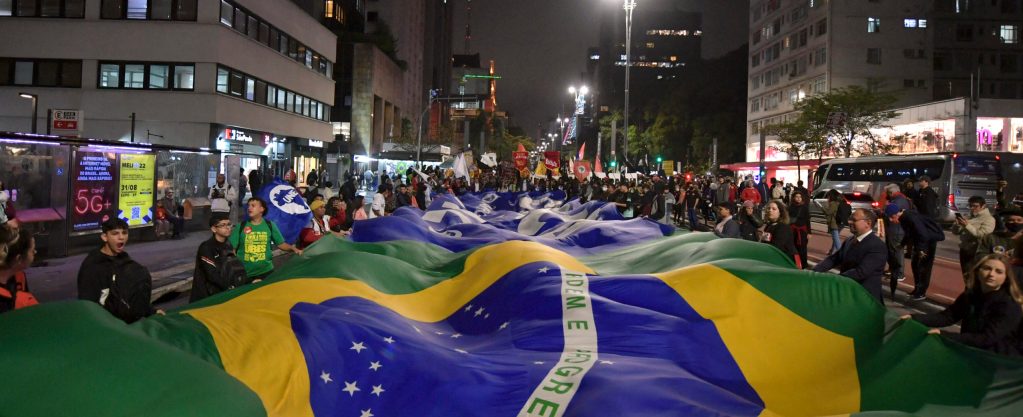 The image shows a crowd of people in front of buildings in the background, apparently holding up a giant green, yellow and blue Brazilian flag in the foreground.