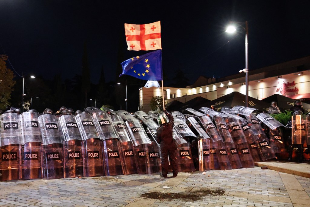 The protester holding the flags stands alone in front of a wall of about 20 officers completely covered by riot shields, each holding two shields vertically.