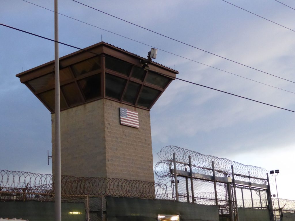 A guard tower is shown with a U.S. flag in the middle and bared wire next to the tower.