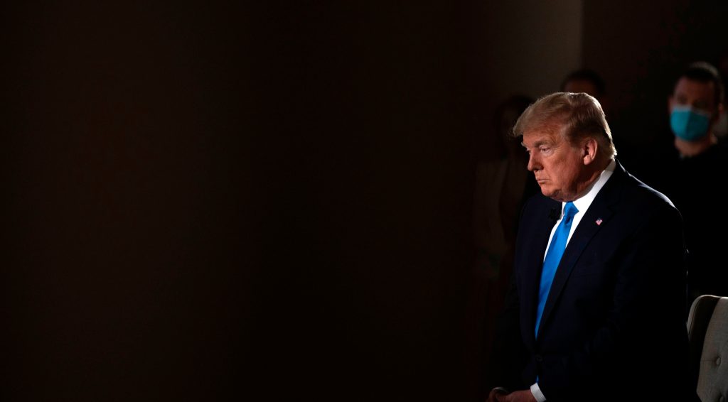 Trump against a dark background. People appear behind him.