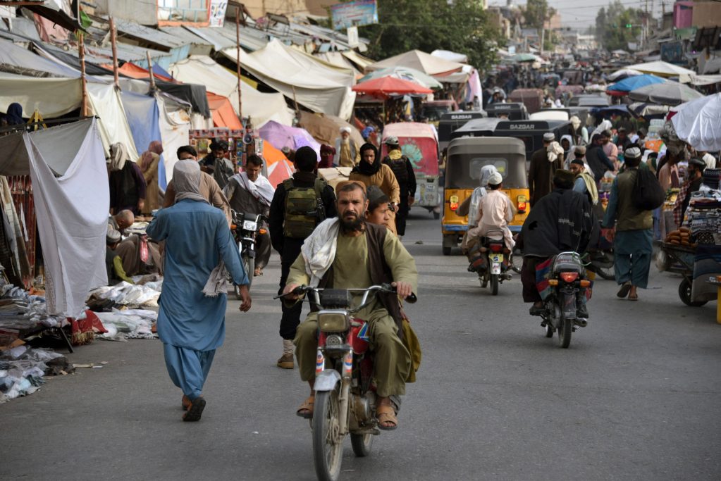 A busy market area in Kandahar. People ride motorized bikes, in small vehicles or walk in the street. Umbrellas and tarps cover market stalls.