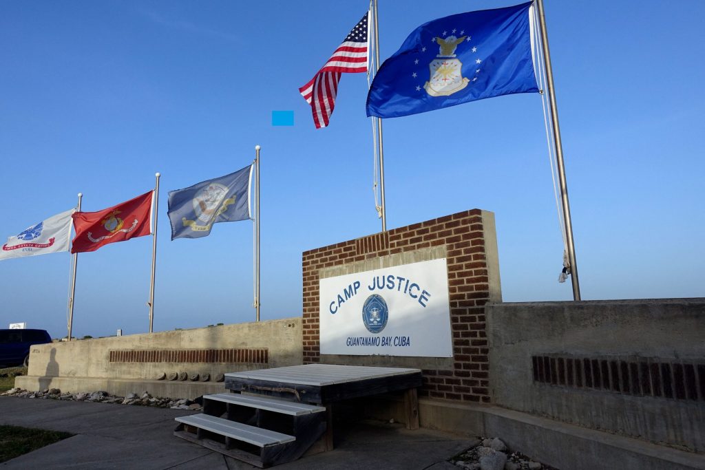 A white sign with blue writing on it reads "Camp Justice" with a blue sky behind it and a U.S. flag flying above.