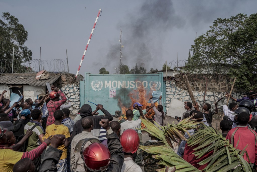 A crowd swirls around a blaze set in front of a blue-green solid metal gate inscribed with the name of the U.N. peacekeeping mission, MONUSCO. A few palm fronds are seen in the foreground, and stone walls flank the gate in the background.