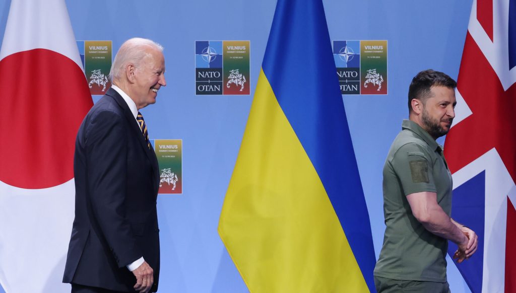 Biden and Zelenskyy walk past a Ukrainian flag. Biden is wearing a dark suit and Zelenskyy is wearing a dark green polo shirt.