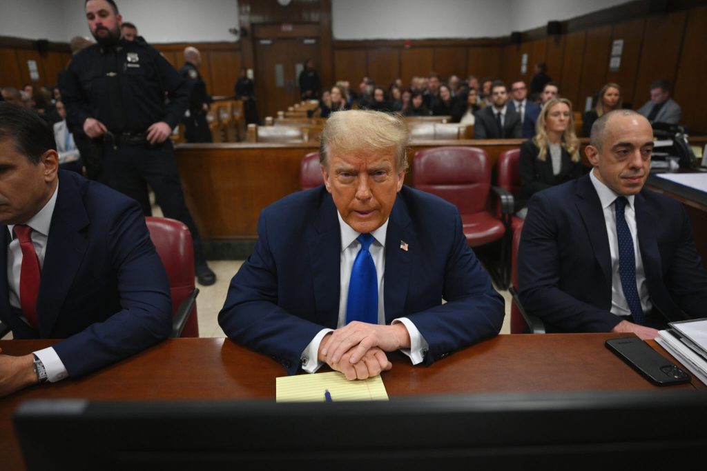 Donald Trump seated at defense table in courtroom, flanked by two lawyers, with public seating in background.
