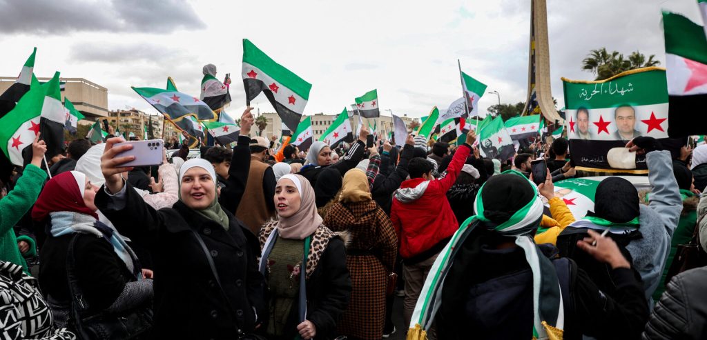 A woman takes a selfie as people wave independence-era Syrian flags during a demonstration