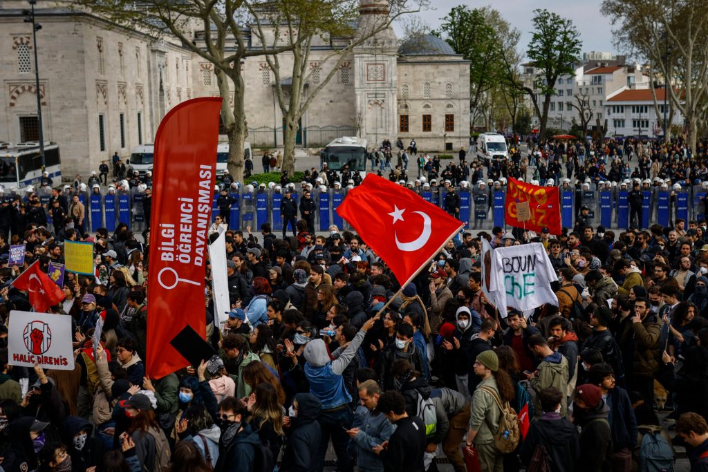 Anti-riot police officers stand guard in a line (Rear) as protesting university students gather. A large Turkish flag sways.