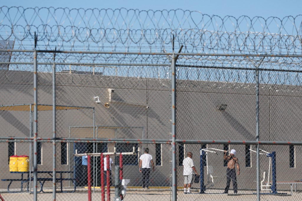 Detained people are standing in front of a building behind a metal, possibly chain link, fence that appears nearly three times taller than they are and topped with concertina wire.