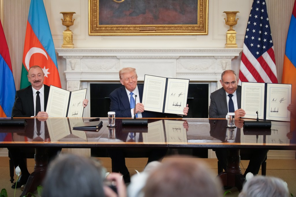 U.S. President Donald Trump (C), Azerbaijani President Ilham Aliyev (L), and Armenian Prime Minister Nikol Pashinyan (R) sit behind a long table, smiling, as they hold up copies of the signed agreement in front of members of the press.