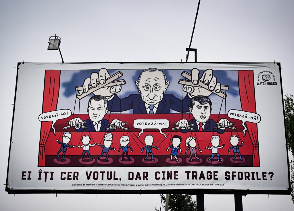 A billboard shows a caricature of Russian President Vladimir Putin pulling the ropes of then-fugitive oligarchs Ilan Shor (R) and Vladimir Plahotniuc (L), as if they are puppets, above words reading "They ask for your vote." The billboard was displayed in Ungheni city on September 24, 2025. (Photo by DANIEL MIHAILESCU/AFP via Getty Images)