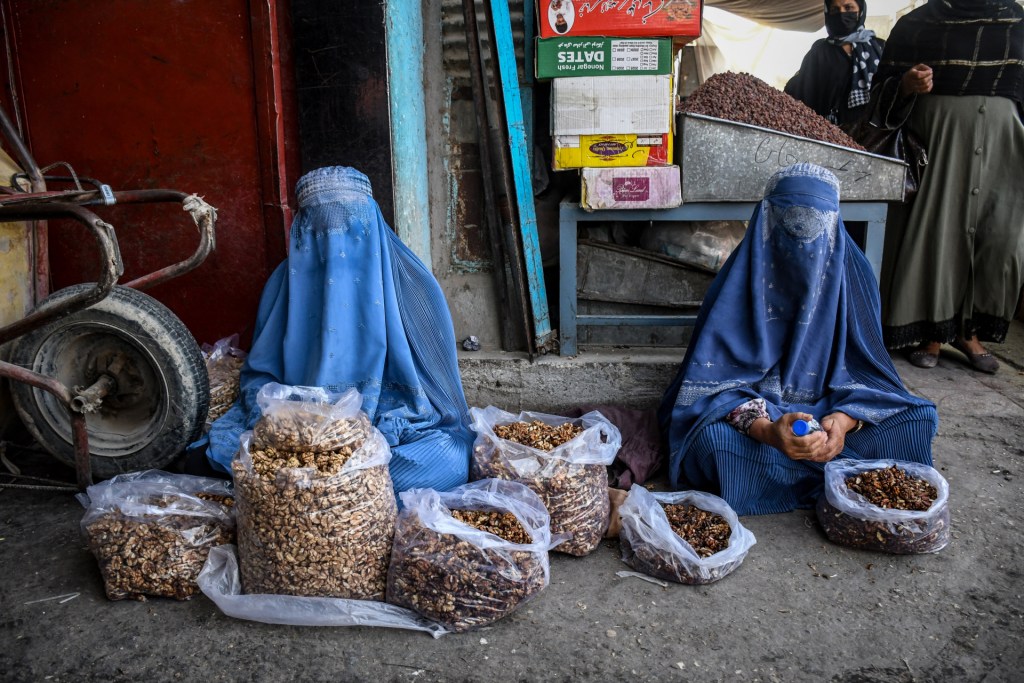 Two Afghan women wearing, from left to right, a light blue and a darker blue burqa sit on the ground with baskets in front of them and what appears to be a more formal market stall behind them, in Mazar-i-Sharif on October 2, 2025. At the left of the photo next to the women is a wheelbarrow turned against a wall. (Photo by ATIF ARYAN/AFP via Getty Images)