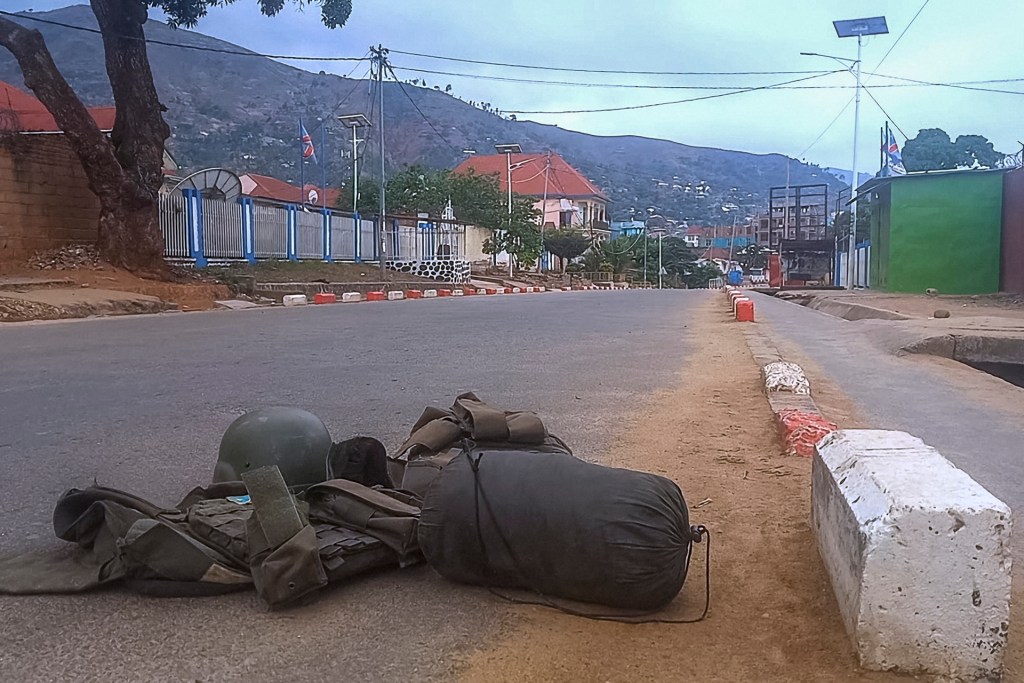 A bullet proof vest carrying a patch with the Democratic Republic of Congo (DRC) flag and a ballistic helmet are lying in an empty road that stretches into the background, alongside other personal belongings. Mountains can be seen stretching across the photo in the background.