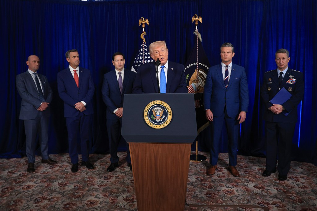 Trump is standing at a wooden lectern with the presidential seal on the front of it. The officials are arrayed behind him, against a dark blue backdrop with an American flag and a presidential flag between the backdrop and the officials. All the men are wearing suits, except Caine, who is wearing a blue Air Force uniform.