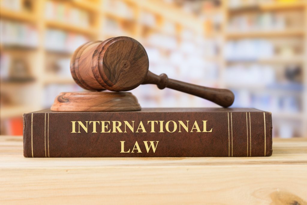 A black colored International Law book sits with a judges gavel on top of it on desk in the library. The book's spine has "International Law" written in gold letters and the gavel is made of dark brown wood.