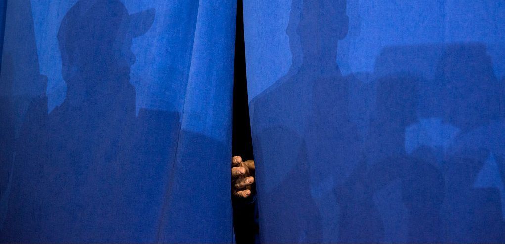 A hand peeks through a blue curtain with shadows of people.