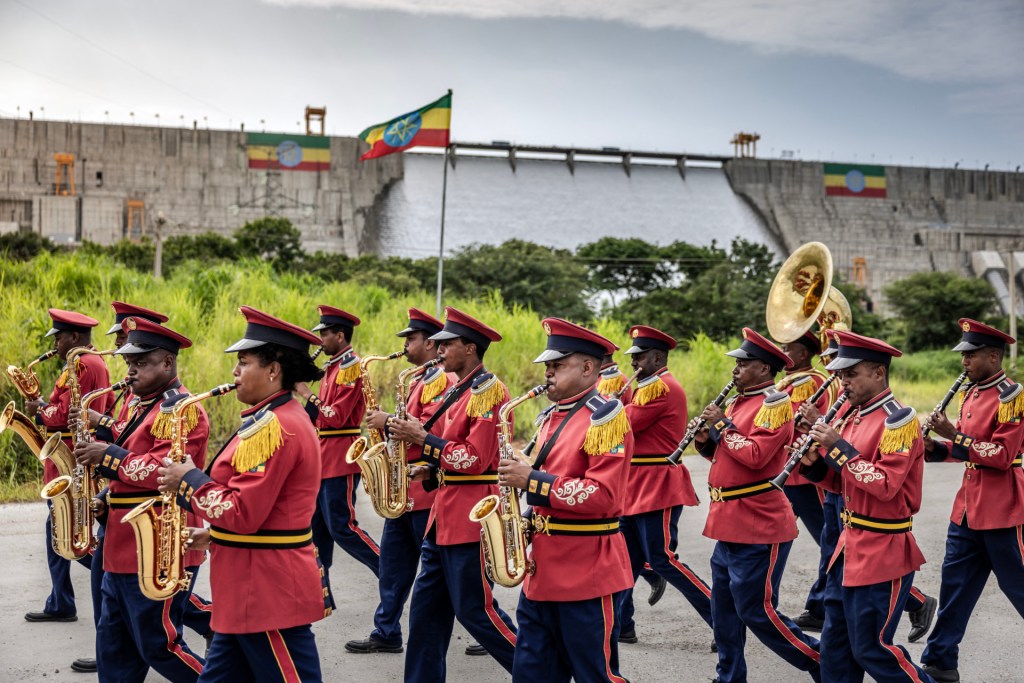 The band members march toward the left of the photo, wearing red jackets and black pants with red stripes and playing instruments including a tuba, as a red, yellow and green Ethiopian flag flies in the background in front of the dam, with water flowing down its slope.
