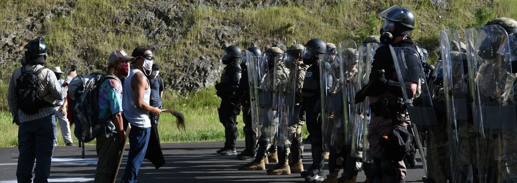 Police and military personnel in full riot gear stand blocking protestors at a demonstration protesting the Mount Rushmore National Monument and the visit of US President Donald Trump.