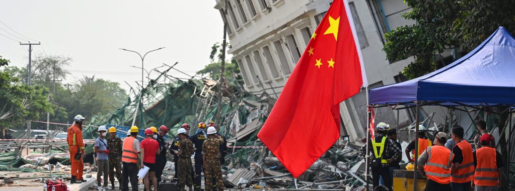 The photo shows a red Chinese flag with gold stars flying over a disaster scene -- a building partly collapsed and leaning, with a pile of rubble at its base and a crowd of rescue workers and civilians around it.