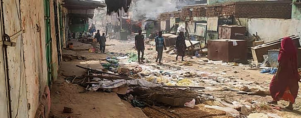 The photo shows three people in the center of the photo walking on barren ground toward the camera, a woman in a red robe and head covering walking toward them at the right of the image, and several people at the back left of the frame. Wooden market stalls on the left edge are closed and covered, and a row of brick and stucco buildings line the right side of the market alley.