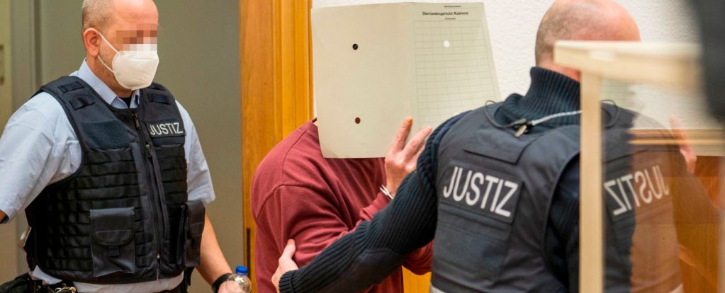 Syrian defendant Eyad al-Gharib, accused of crimes against humanity in the first trial of its kind to emerge from the Syrian conflict, arrives to hear his verdict in the court room on February 24, 2021 in Koblenz, western Germany. He holds up a document or folder to hide his face as two officers in bullet proof vests touch him and move him forward.