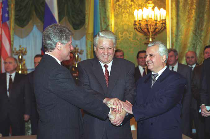 U.S. President Clinton, Russian President Yeltsin, and Ukrainian President Kravchuk engage in a three-way handshake against a backdrop of a richly decorated room with fringed drapes and a chandelier-like wall sconce in the background.